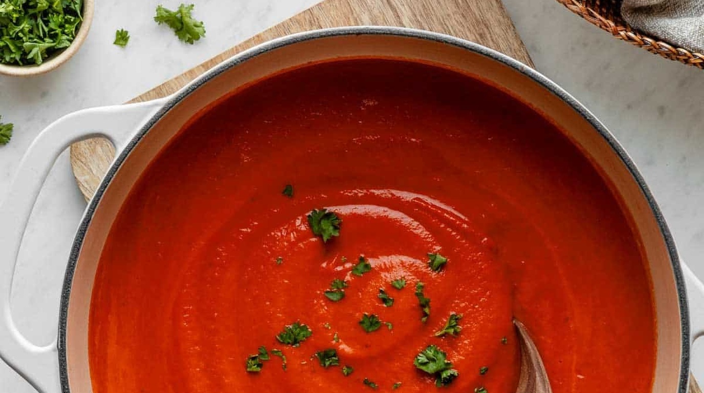 Ultra close-up macro of velvety orange-red creamy roasted tomato basil soup swirling with heavy cream and fresh green basil