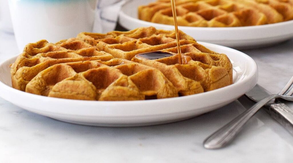 Ultra close-up macro of golden crispy pumpkin waffles with warm maple syrup dripping into deep pockets