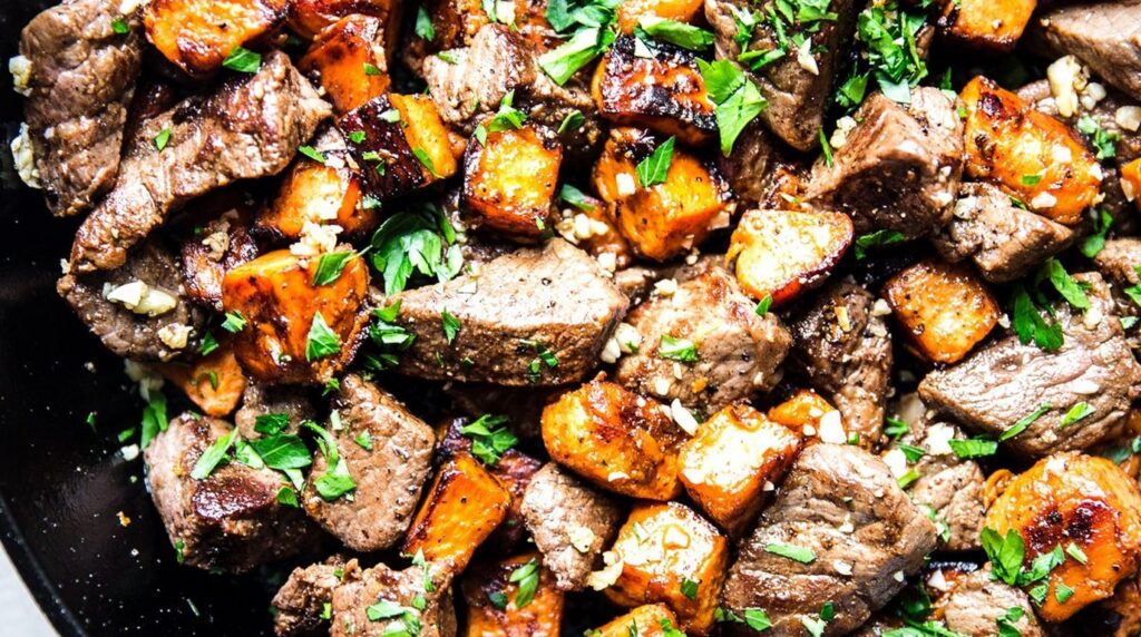 Ultra close-up macro of sizzling garlic butter steak bites and caramelized sweet potatoes in a cast-iron skillet